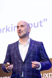 A male speaker on stage wearing a purple suit and presenting a talk